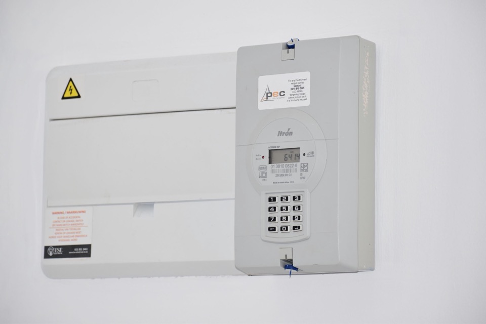 Een meterkast op een witte muur met een kWh-meter en een gele waarschuwingssticker