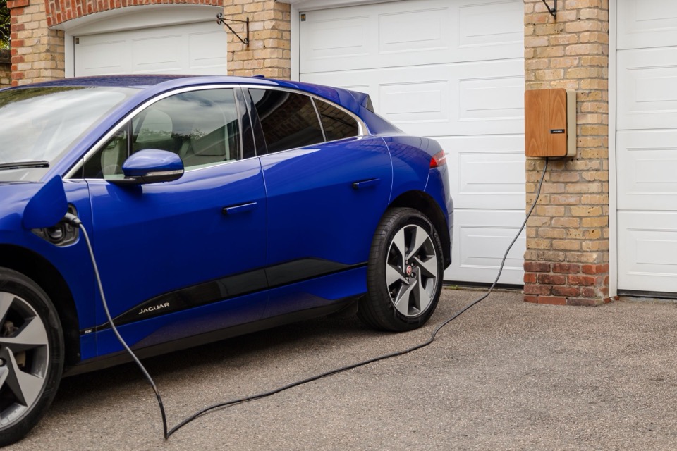 Een blauwe elektrische auto laadt voor een garage met een houten kastje aan de gevel